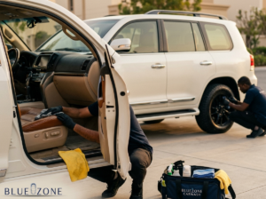 Two car wash workers detail a white SUV in a driveway: one kneels inside the open door to wipe the tan leather seat, the other crouches by the rear wheel; a BlueZone Carwash kit sits on the ground nearby.