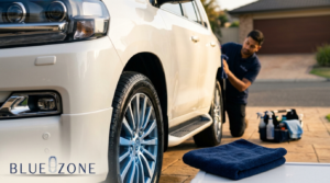 Car being detailed in a driveway with cleaning supplies, a technician wiping the vehicle’s side panel beside a folded towel.