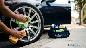 Person wiping a chrome alloy wheel with a green cloth, car door open in the background and a BlueZone carwash kit nearby.