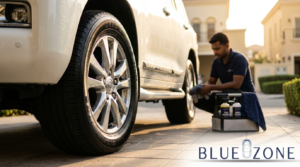 Tech kneeling by a white SUV, hand polishing the wheel with a detailing kit on a driveway, Blue Zone logo visible in corner.